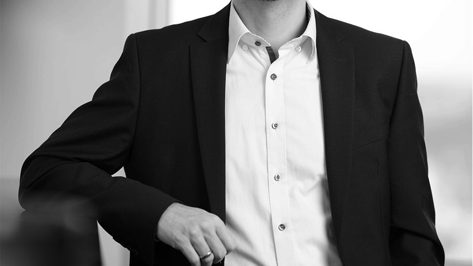 Das Bild zeigt einen Mann in formeller Kleidung, bestehend aus einem dunklen Anzug und einem hellen Hemd. Er steht aufrecht und hält mit einer Hand eine nicht sichtbare Kante oder einen Gegenstand. Der Hintergrund ist unscharf, wodurch der Fokus auf die Person gerichtet ist.